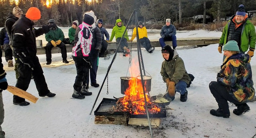 a group of people wearing winter gear stand around a campfire with two pots hanging from a tripod over the flames